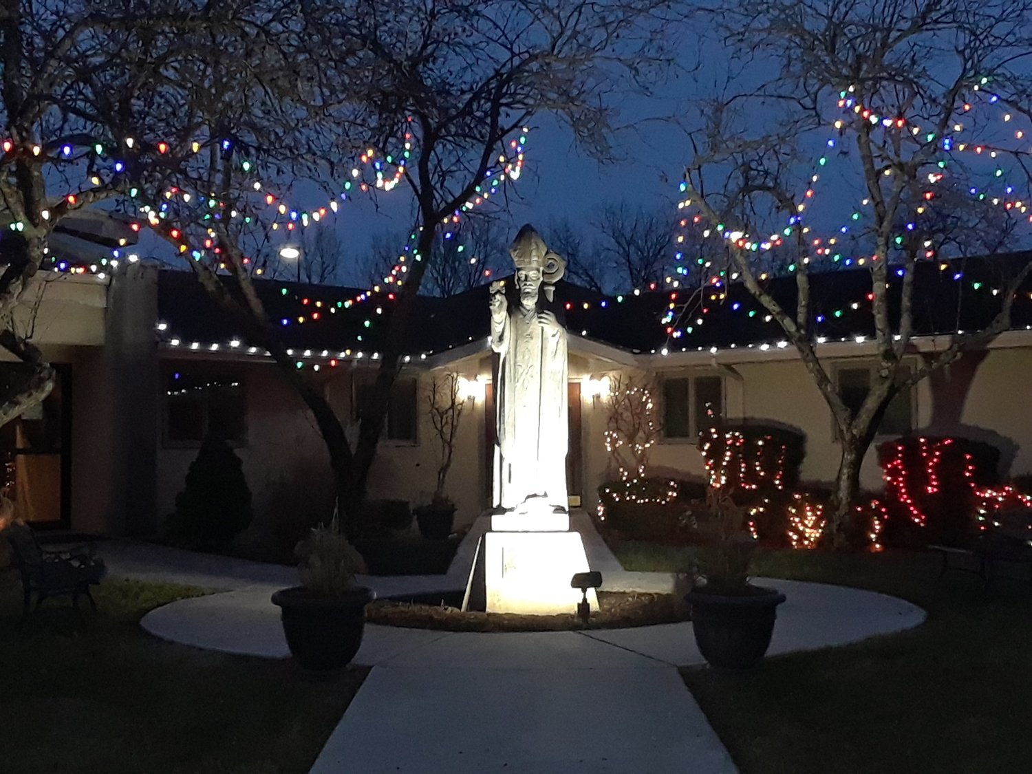 Christmas lights adorn the wedge-shaped garden between St. Patrick Church and the parish rectory in Laurie, where this limestone image of St. Patrick arrived providentially on St. Patrick’s Day in 1980.